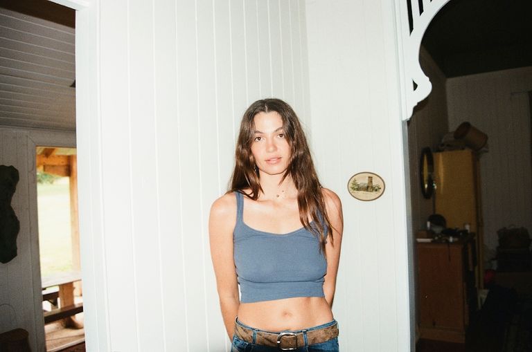 Australian singer-songwriter Kita Alexander wearing a blue tank top and jeans, standing against a white paneled wall inside a home.